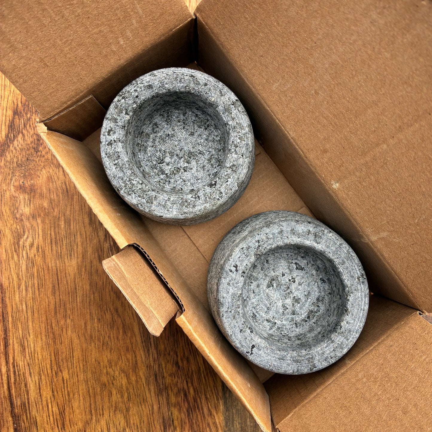 Two grey stone bowls in a cardboard box on a wooden surface