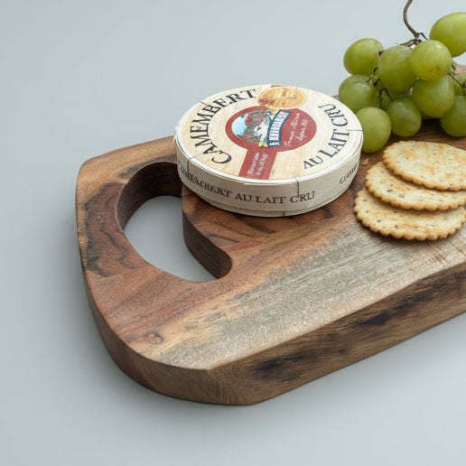 close up image of a Wooden cutting board with a wheel of cheese, crackers, and grapes on a gray surface