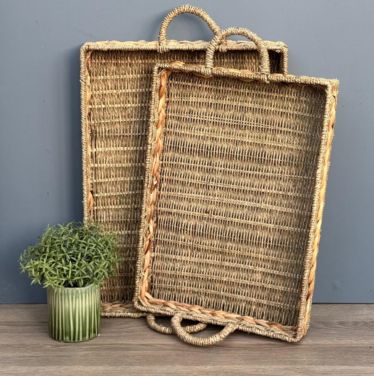 Two woven trays with handles on a wooden surface against a gray wall.