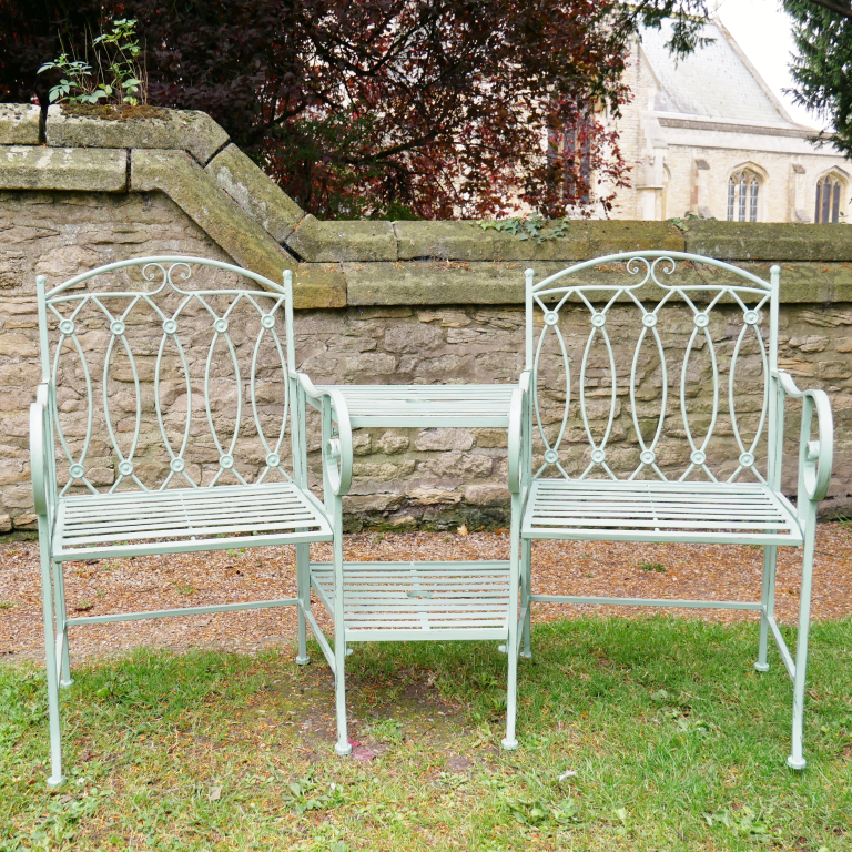 Two metal chairs with decorative backrests on a grassy area near a stone wall.