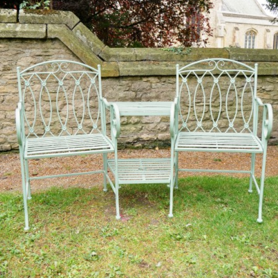 Two white metal chairs on a grassy area with a wooden fence and trees in the background.