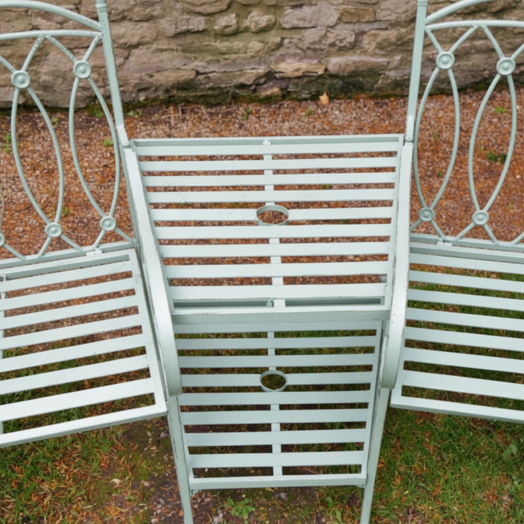 Wrought iron garden bench with decorative backrest on grass against a stone wall.