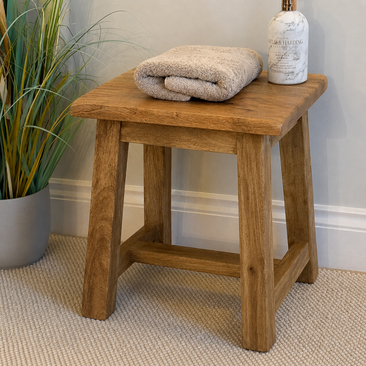 A rustic wooden bath stool with a natural oak finish, styled with a folded beige towel and a white bottle on top, set in a bathroom corner next to a potted plant.