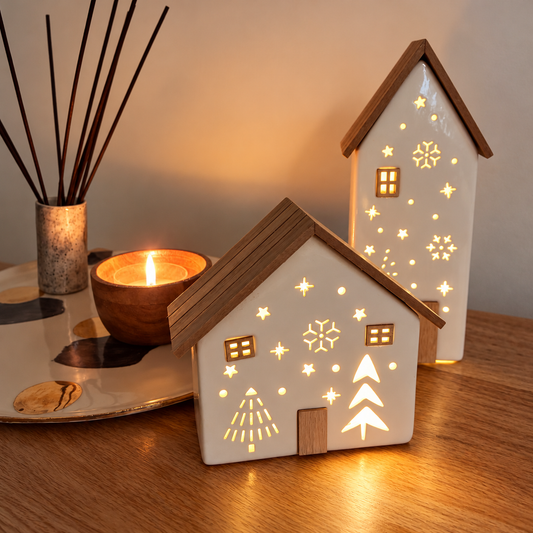 Two small house-shaped ceramic night lights with warm white glow and star, snowflake, and tree cutout designs, displayed on a wooden table next to a lit candle and reed diffuser.