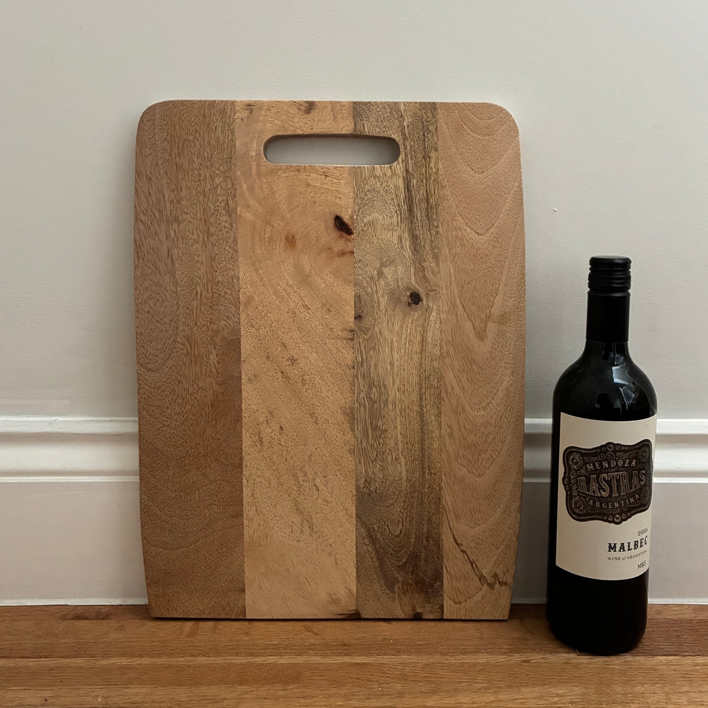 Wooden cutting board with a bottle of wine on a wooden surface against a neutral wall.