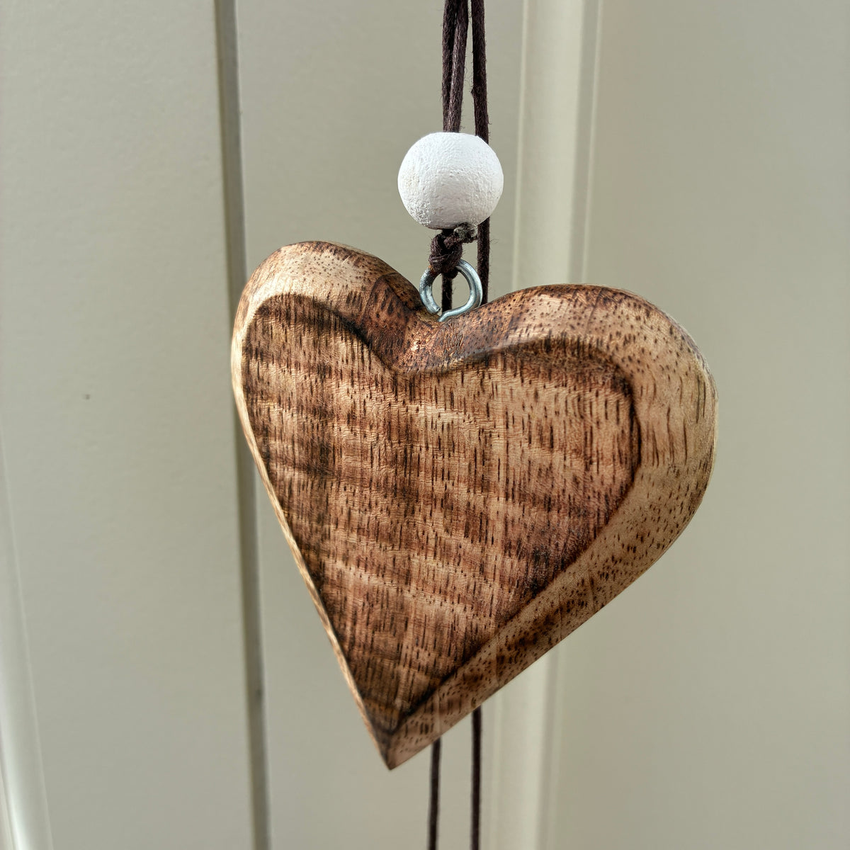 A rustic, hand-carved wooden heart ornament with a dark brown and tan wood grain pattern, hanging from a dark brown cord with a white bead.