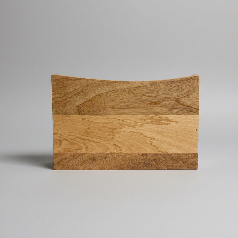A rectangular wooden board or block with a curved top edge, made from three horizontal bands of natural wood grain, standing upright on a light-colored surface against a dark gray background.