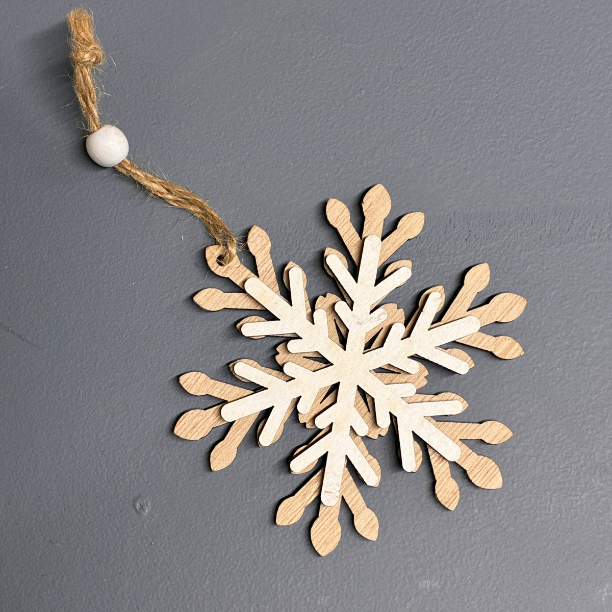 A wooden snowflake ornament with layered light natural wood cutouts, hanging from a tan jute string and a small white bead, shown on a dark gray background.