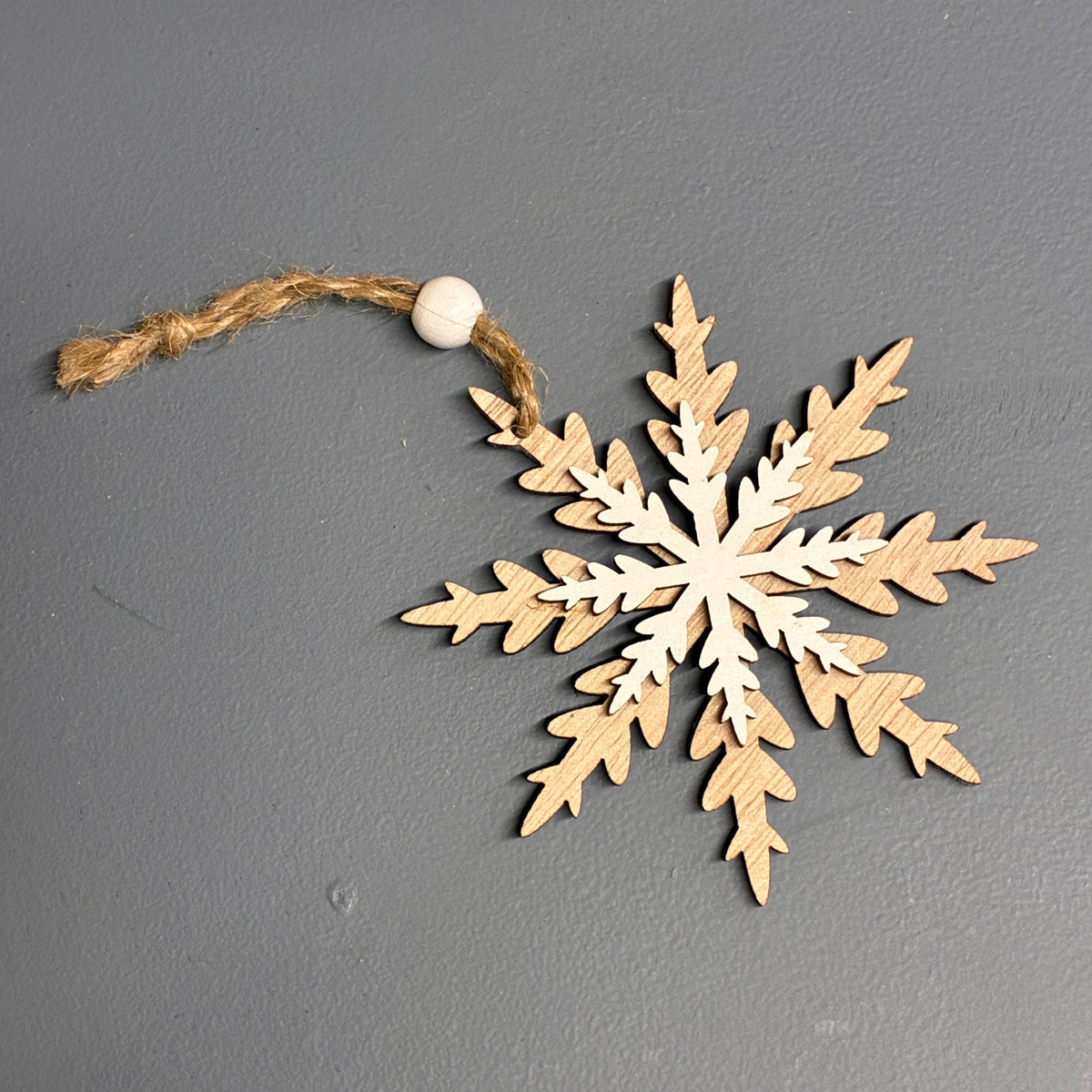 A natural wood snowflake hanging ornament with a jute rope hanger and a small white bead, displayed on a dark gray textured background.