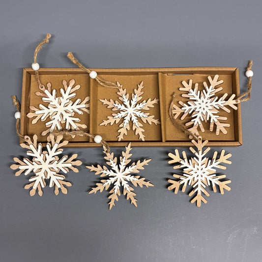 A set of six wooden snowflake ornaments in natural light wood, displayed in a brown cardboard tray with three hanging above and three resting in front, against a solid dark gray background.