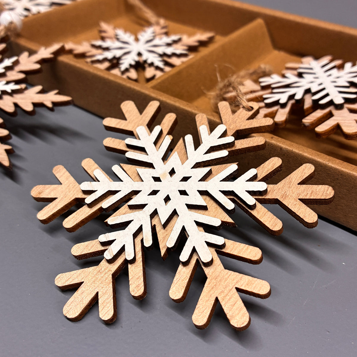 Laser-cut wooden snowflake ornaments in natural light brown wood with white inner cutouts, displayed on a dark gray surface with a cardboard tray of more ornaments in the background.