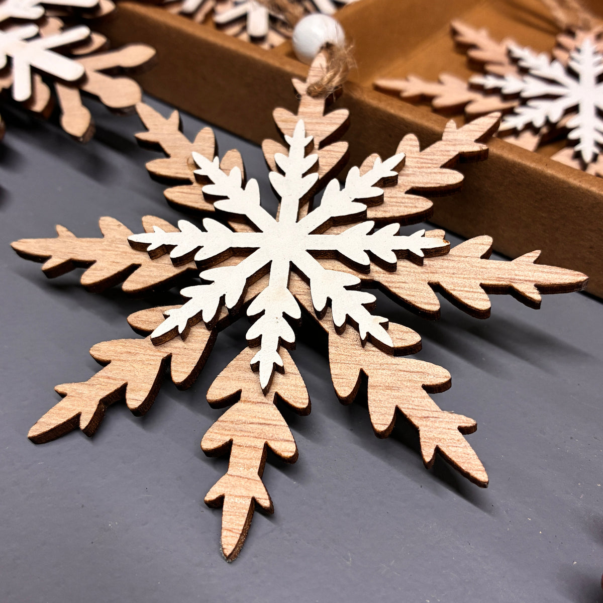 A close-up of a wooden snowflake ornament layered over a white snowflake cutout, with additional ornaments and a brown box in the background.
