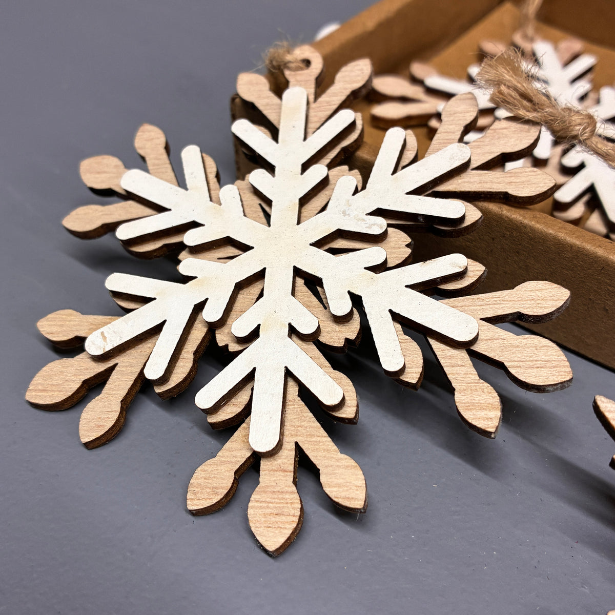 Unpainted wooden snowflake ornaments stacked on a dark gray surface, with a brown cardboard tray holding more ornaments in the background.