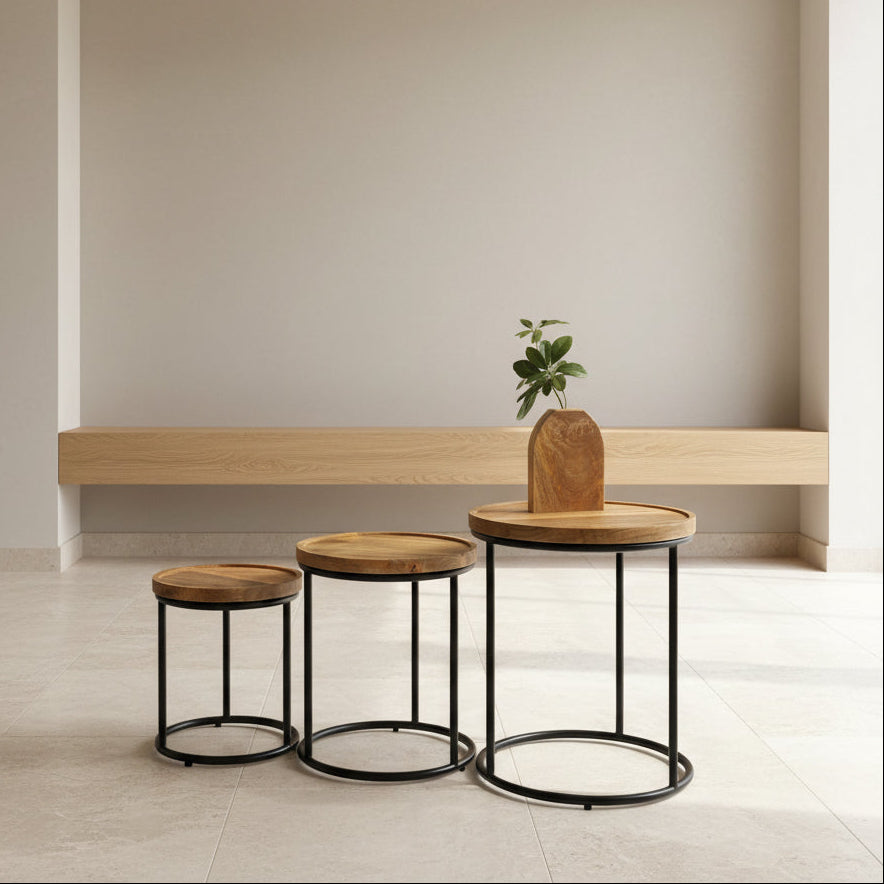 Three round wooden side tables with black metal circular pedestal bases, arranged on a white-covered table in a warehouse setting. A small vase with a green plant sits on the largest table.
