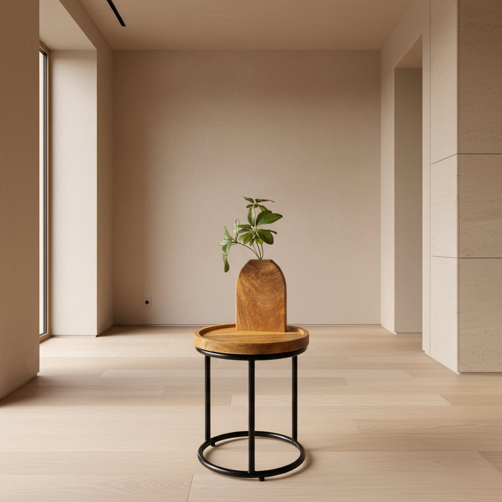 A small round wooden side table with a black metal circular stand, holding a carved wooden planter with a green plant, set against a gray backdrop.