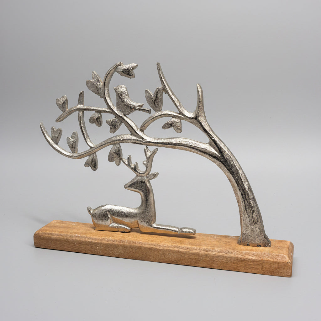 A decorative metal sculpture of a deer lying beneath a tree with birds, mounted on a rectangular wooden base against a dark gray background.