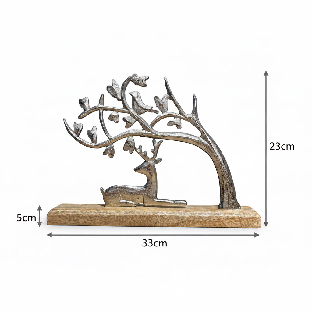A decorative metal sculpture of a deer beneath a tree with birds, mounted on a rectangular wooden base, photographed against a plain blue-gray wall and tan surface.