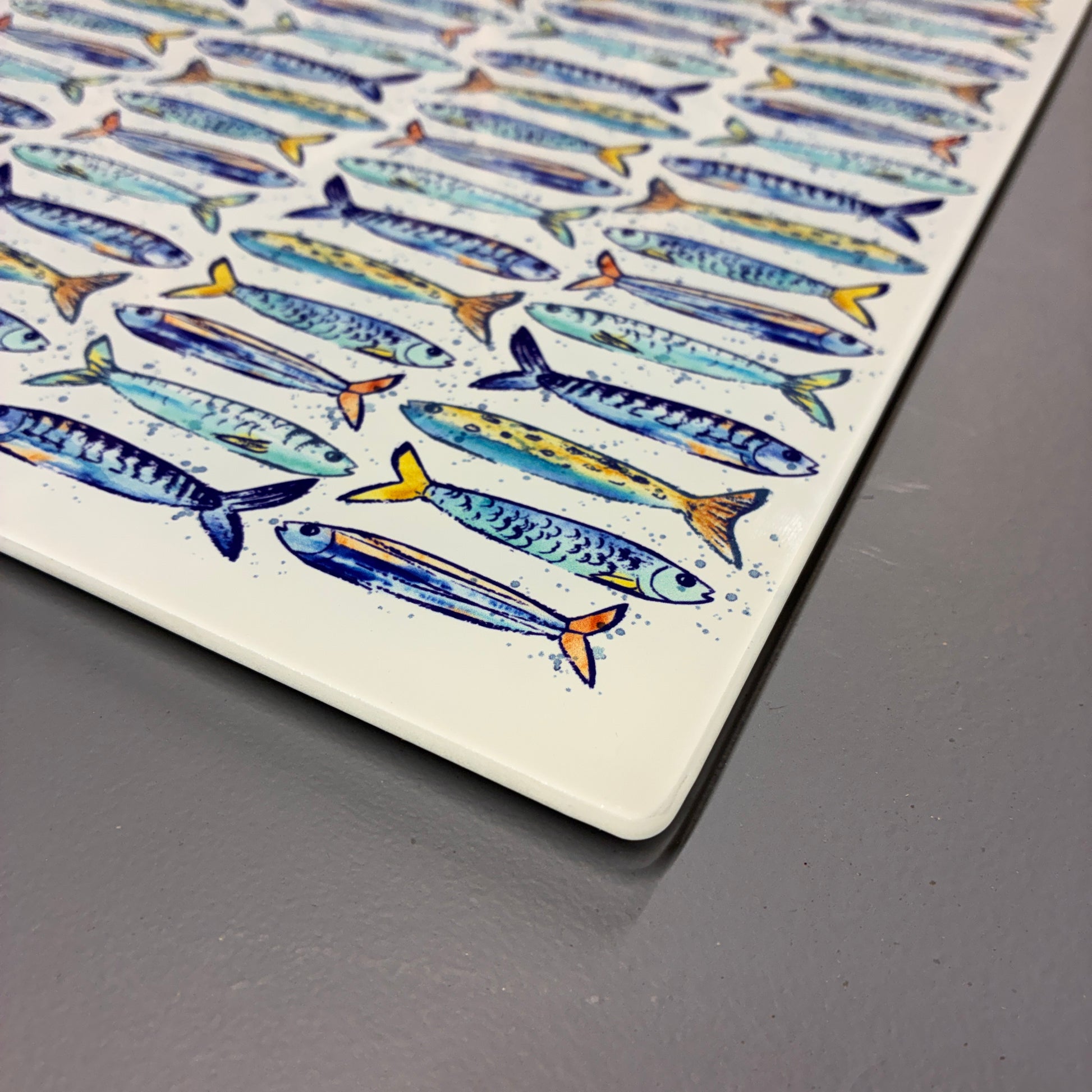 A square ceramic plate with rounded corners, featuring a repeating pattern of illustrated blue, yellow, and orange fish on a white background, shown resting on a dark gray surface.