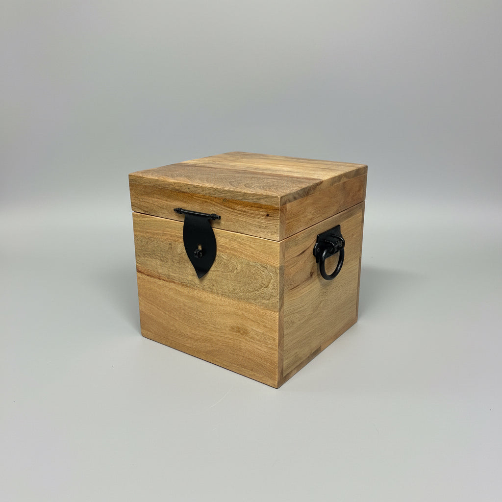A square wooden storage box with a light brown wood grain finish and black metal hardware, including a central strap clasp and side rings, resting on a white textured surface in a workshop setting.
