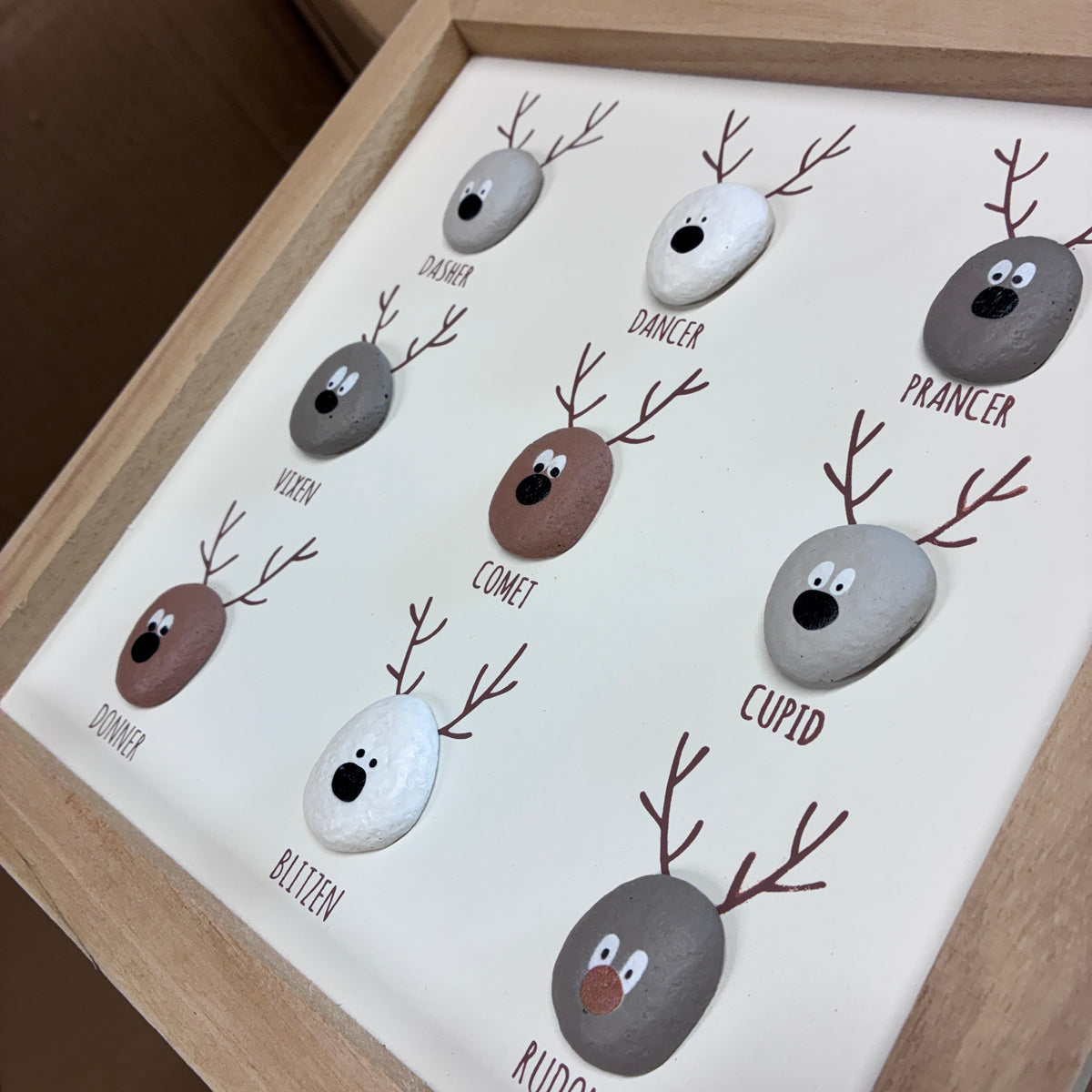 A framed decorative display featuring nine small painted stone heads with antlers and names such as Prancer, Viten, Comet, Cupid, Blitzen, and Rudolph. The stones are arranged on a white backing inside a light wood frame.