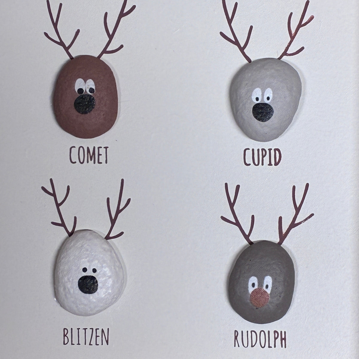 Four hand-painted decorative stones arranged in a 2x2 grid on a white background, each styled as a reindeer head with brown antlers and labeled COMET, CUPID, BLITZEN, and RUDOLPH.