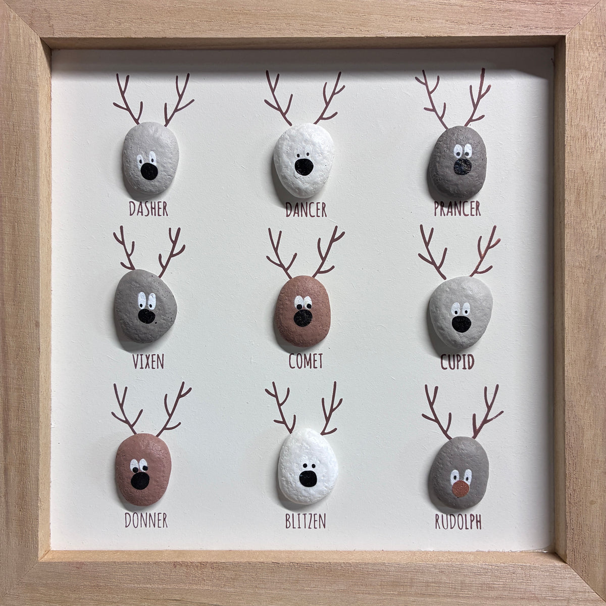 A framed 3x3 custom pet memorial art print featuring hand-painted river stones shaped like reindeer heads, each labeled with a different pet name: Dasher, Dancer, Pointer, Vixen, Comet, Lupus, Donner, Blitzen, and Rudolph. The stones are arranged in a grid on a white background inside a light wood frame.