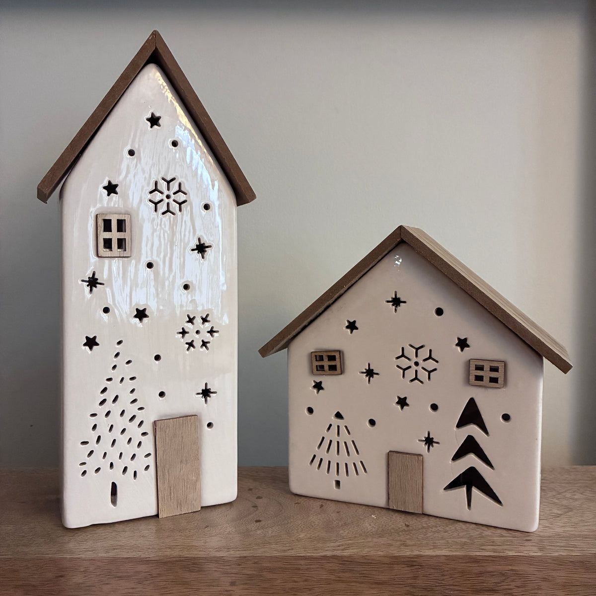 Two small decorative house-shaped ornaments or figurines standing on a wooden surface against a plain light gray wall. The larger one on the left is glossy white with brown trim and black star, snowflake, and tree cutout designs.