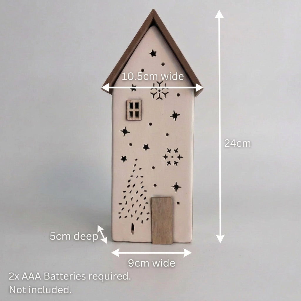 A small decorative house-shaped ornament with a white body, dark brown roof and door, and black star and snowflake cutouts, standing on a wooden surface against a plain light gray wall.
