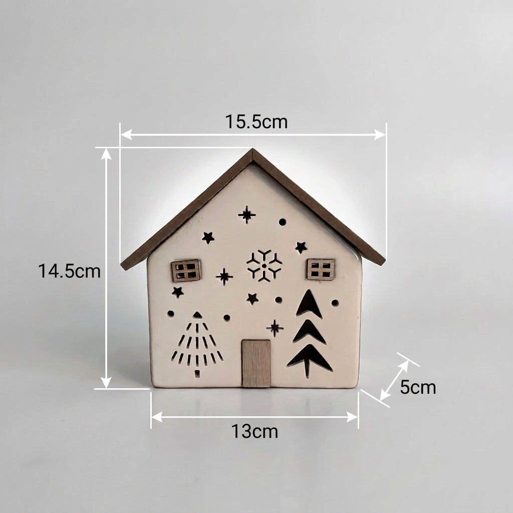 A small decorative house-shaped ornament or candle holder with a white body, brown roof and trim, black star and tree cutouts, and two small square window details, displayed on a wooden surface against a plain light gray wall.