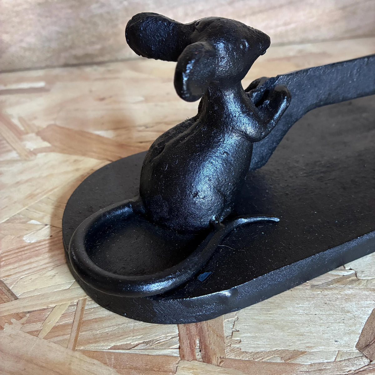 A black cast iron fireplace poker with a sculpted mouse figure on the handle, resting on a light wood particle board surface.