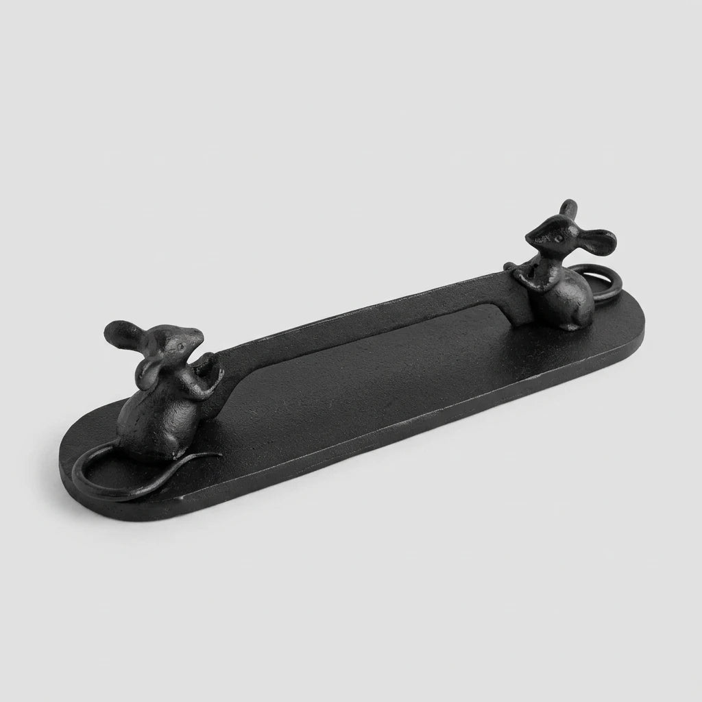 A black cast iron mouse-shaped tray with two small mouse figures at the ends, photographed on a light wood particle board surface.